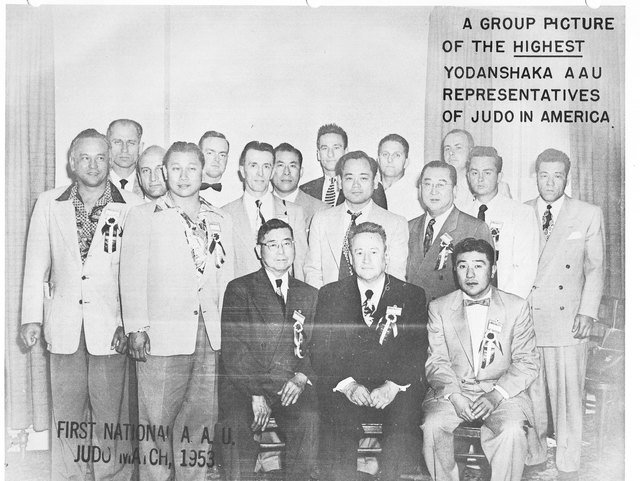 A black and white group photograph of eighteen men in suits, three seated and the rest standing in rows, before a pale curtain. A banner behind them reads: a group picture of the highest yodanshaka AAU representatives of judo in America.