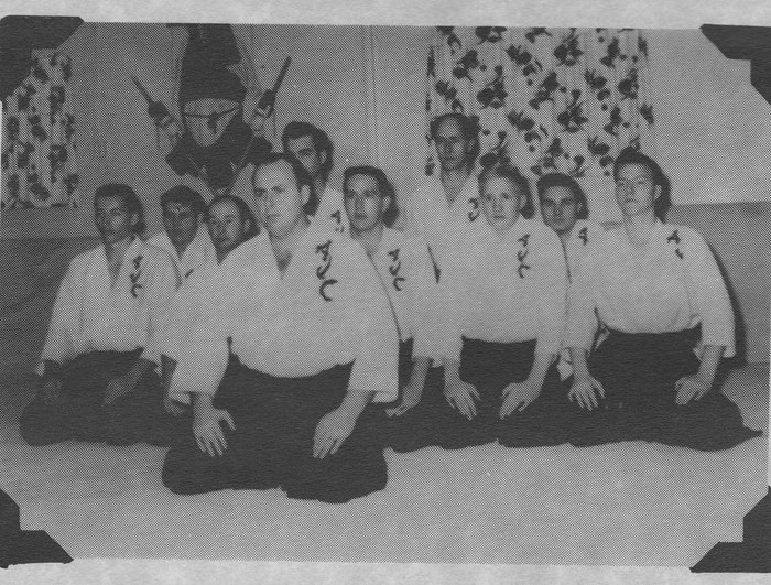 A black and white photograph of nine Western men kneeling in two rows on a mat, in white gi lettered AJC in Japanese characters and dark hakama, before a decorated wall.