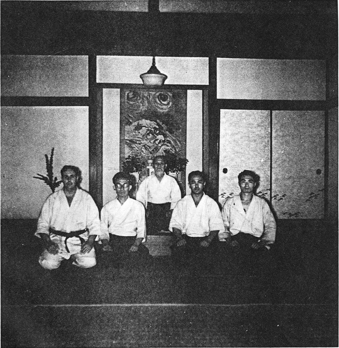 A black and white photograph of five men kneeling in a row on tatami before a lit shrine alcove with a painted panel above it. The man on the left is Western; the bearded figure at the centre kneels furthest back, before the shrine.