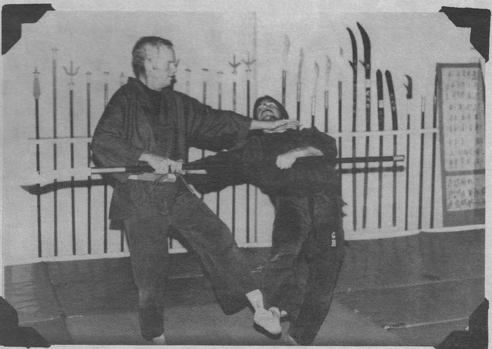 A black and white photograph in a dojo hung with polearms and swords. An older man in a dark gi has stepped inside a spear thrust and is taking the weapon away from a younger man, whose head is driven back.