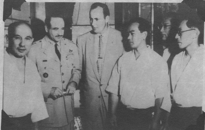 A black and white photograph of five men standing indoors. Second from left a uniformed US Army general with decorations; beside him a tall Western man in a light suit; to the right two Japanese men in white shirts.