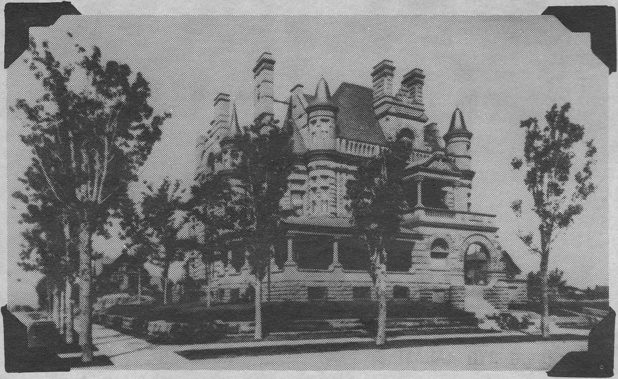 A black and white photograph of a large Victorian mansion behind bare young trees: turrets with conical roofs, tall chimneys, a stone arch over the carriage entrance and a broad flight of steps to the front door.