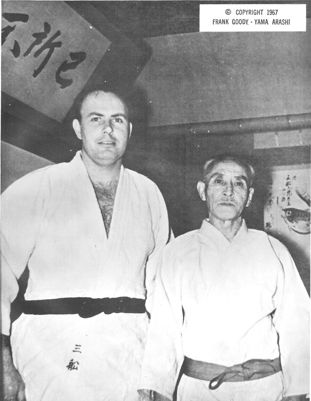 A black and white photograph of two men in judogi before a Japanese dojo wall hung with calligraphy. On the left a tall young Western man with a black belt, two Japanese characters brushed low on his jacket; on the right a small elderly Japanese man with a worn belt.