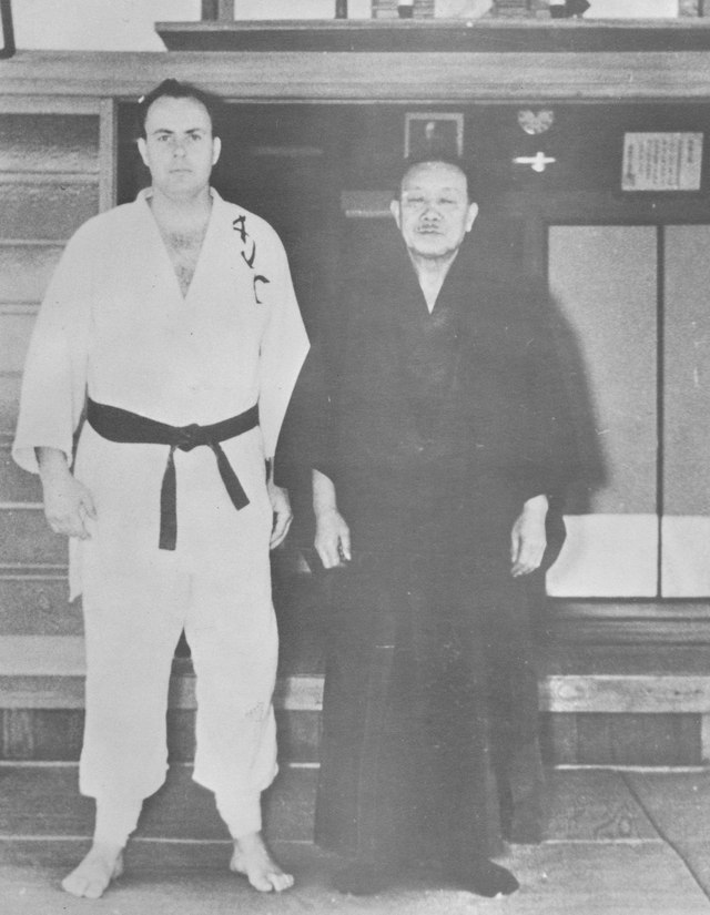 Frank Goody in a judogi lettered AJC standing beside Nagai Sensei in dark formal dress, on the step of a Japanese dojo, 1965.