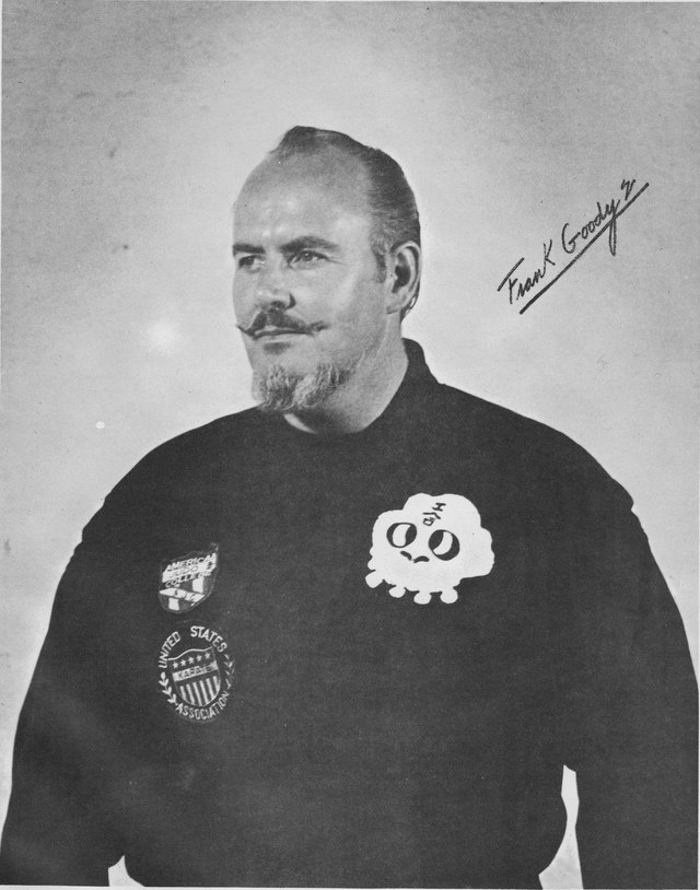 A studio portrait of Frank Goody in a dark gi, heavy-set, with a moustache and goatee and receding hair, two embroidered badges on the chest. Signed across the upper right in white ink.