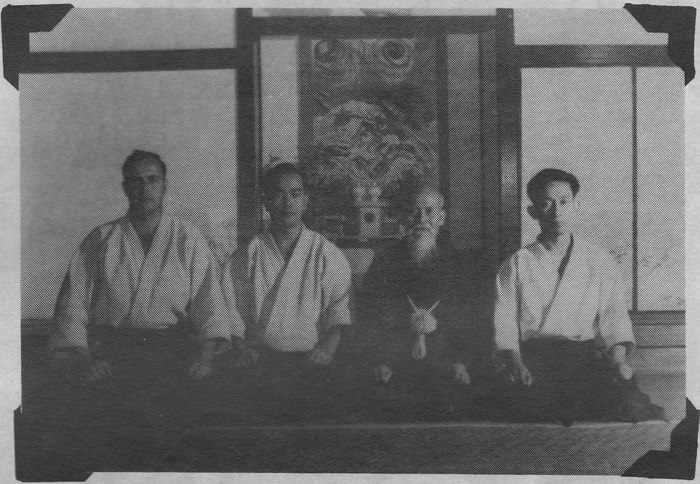 A black and white photograph of four men kneeling in seiza on tatami before a dojo shrine hung with a painted panel. On the left a Western man in keikogi; beside him a Japanese man in glasses; then an elderly man with a full white beard; then a younger Japanese man.