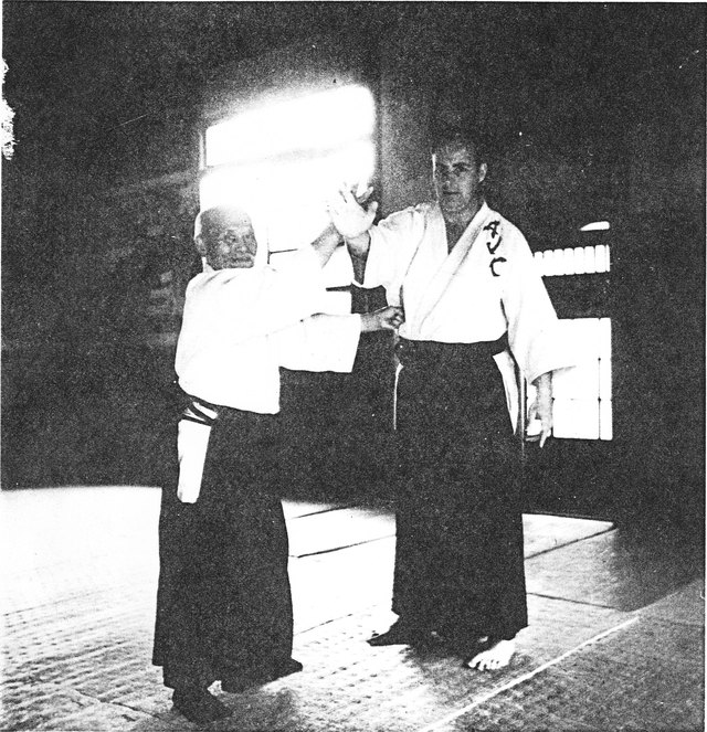 A black and white photograph taken on a dojo mat. A small elderly Japanese man in white gi and dark hakama has hold of the wrist of a much taller Western man in a gi lettered AJC, who is off balance.