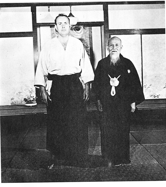 A black and white photograph of two men standing on a stone step before the shoji screens of a Japanese dojo. On the left a tall Western man in white keikogi and dark hakama; on the right an elderly Japanese man, bald with a full white beard, in a dark crested kimono with a tasselled cord.