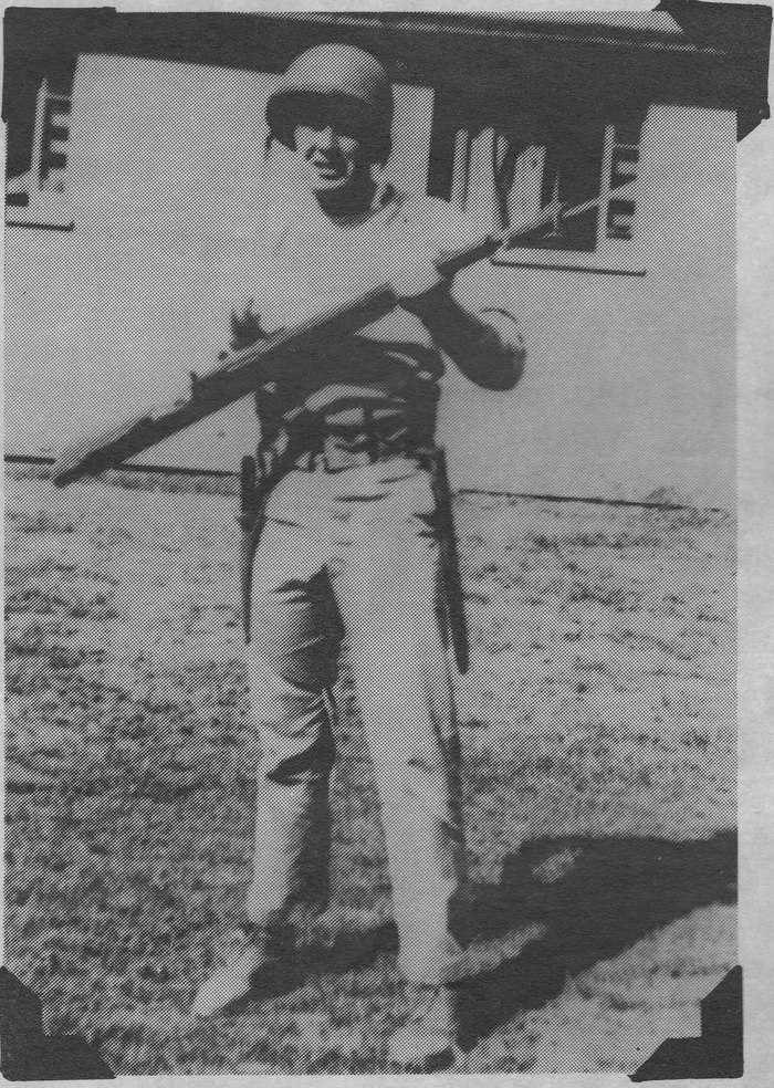 A black and white photograph of a soldier in helmet and fatigues standing before a wooden building, a rifle with fixed bayonet held horizontally across his shoulders.