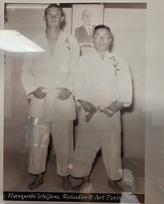 A framed black and white photograph hanging in the dojo office: two men in judogi lettered AJC standing side by side. On the left a tall fair-haired Western man, Art Doris; on the right a shorter man in glasses, Haruyoshi Ichijima. A framed portrait of an elderly man in a suit hangs behind them. A caption band beneath reads Haruyoshi Ichijima, Rokudan and Art Doris, 1957.
