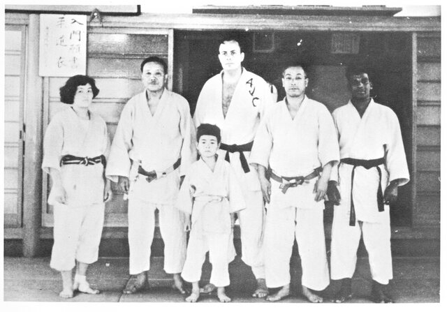 Six people outside a wooden dojo in Tokyo, 1965: five adults in judogi and a boy. A Western man stands tallest at the centre, his jacket lettered AJC.