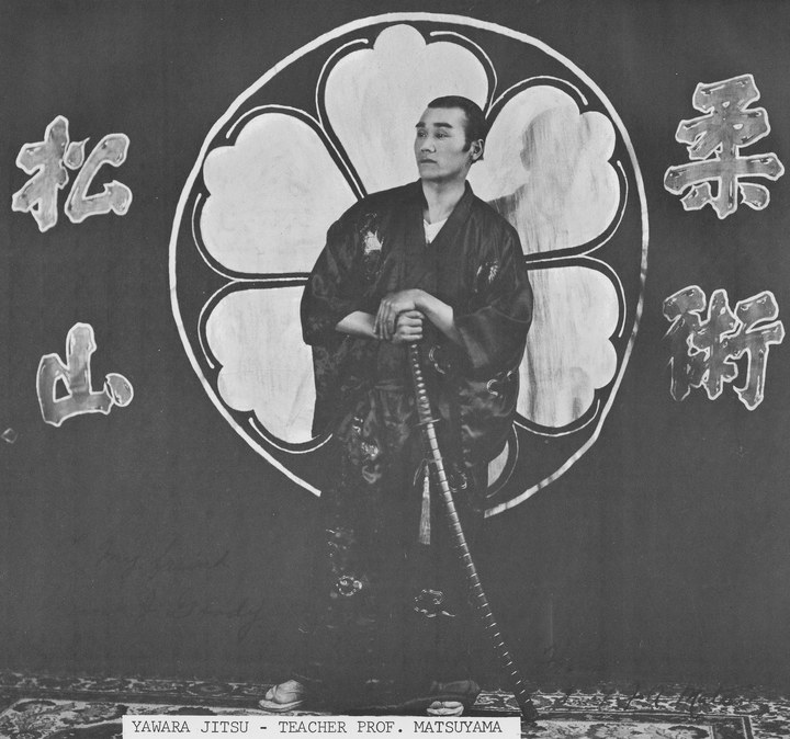 Professor Matsuyama in a dark crested kimono, holding a sword with its tip on the floor, before a large painted crest on a dark drape with brushed characters either side of him.