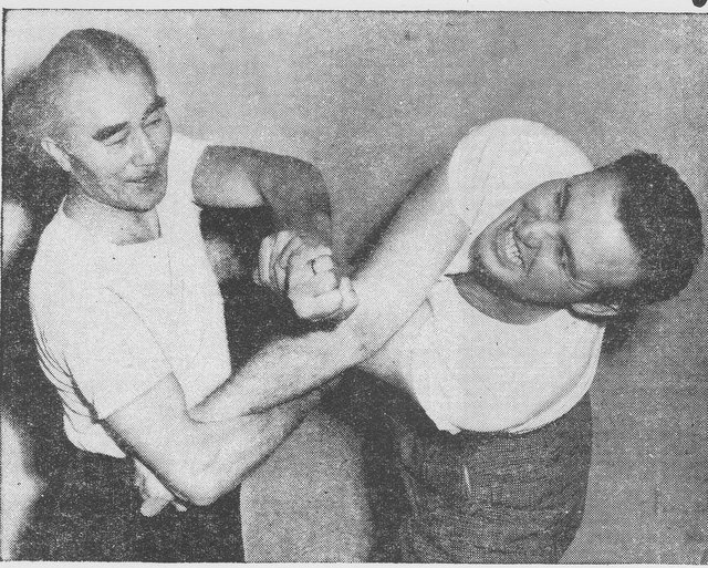 A black and white press photograph of two men in shirtsleeves. On the left an older man with glasses and greying hair has trapped the punching arm of a younger, heavier man on the right, who is grimacing and off balance.