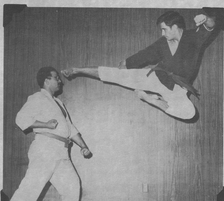 A black and white photograph of two men in a dojo. One stands braced in a gi holding a microphone; the other is completely airborne beside him, body horizontal, driving a side kick out at the standing man's head height.