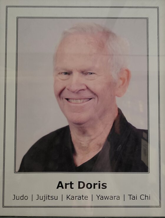 A framed portrait hanging in the dojo office: a smiling older man with close-cropped fair hair, in a dark patterned gi. The mount is captioned Art Doris, and beneath that Judo, Jujitsu, Karate, Yawara, Tai Chi.