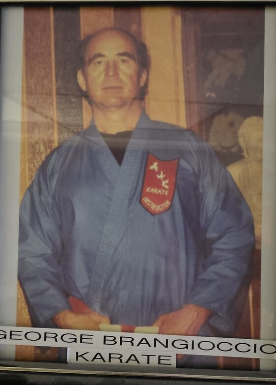 A framed colour portrait hanging in the dojo office: a man in a blue gi with a red shield patch on the chest and a dark belt. A label beneath reads George Brangioccio, Karate.