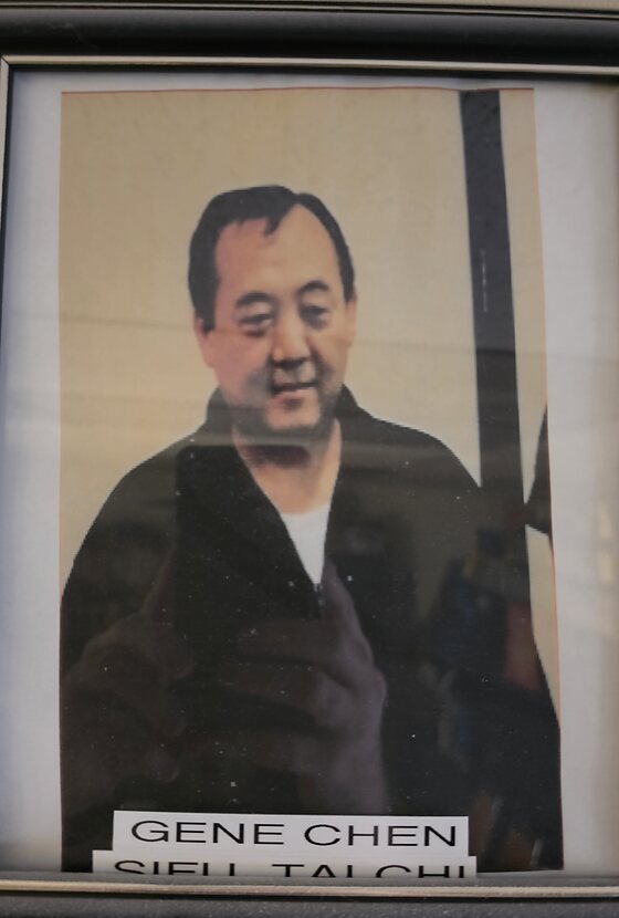 A framed portrait hanging in the dojo office: a man with dark hair in a dark jacket over a white shirt. A label beneath reads Gene Chen, Sifu Tai Chi.