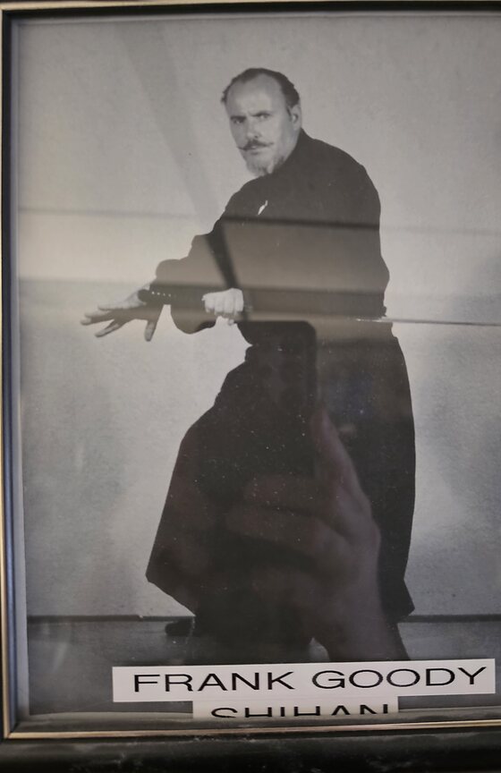 A framed black and white photograph hanging in the dojo office: a bearded man in a dark gi, arms extended in a kata posture against a plain wall. A label beneath reads Frank Goody, Shihan.