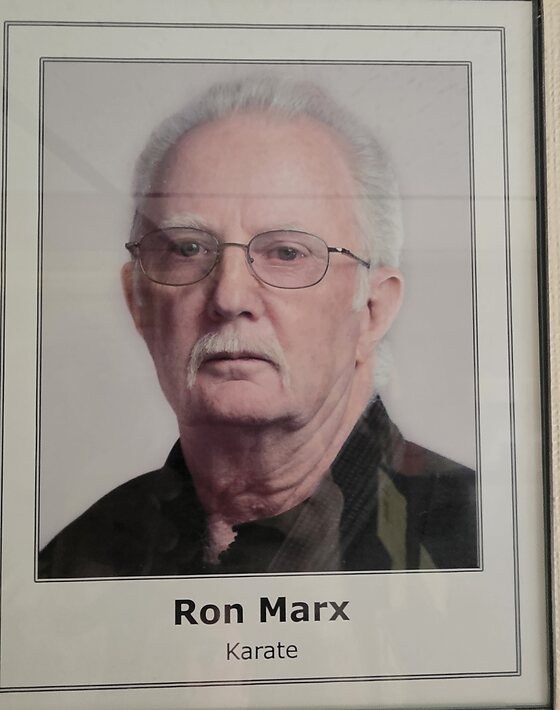 A framed portrait hanging in the dojo office: a white-haired man in glasses and a white moustache, wearing a dark gi. The mount is captioned Ron Marx, Karate.
