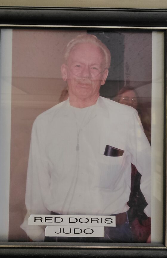 A framed portrait hanging in the dojo office: an elderly man in a white jacket with a dark pen case in the breast pocket. A label beneath reads Red Doris, Judo.